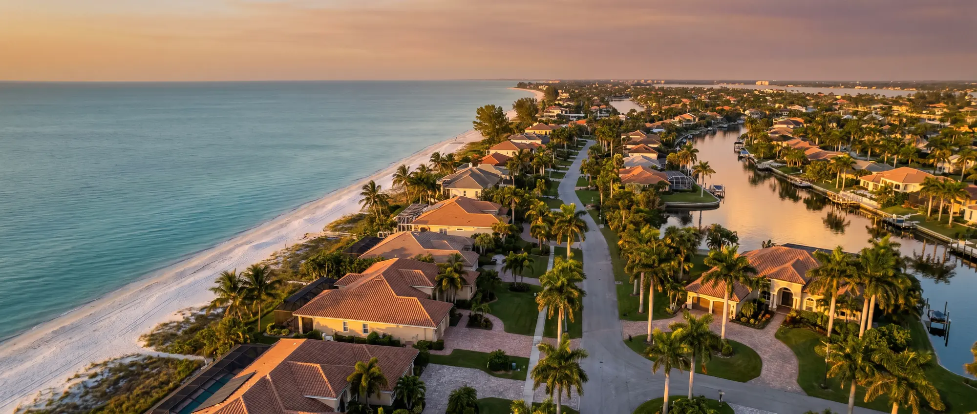 Aerial view at golden hour of a Southwest Florida gulf coast neighborhood: white sand beach and turquoise water on the left, canals and single-story homes with terracotta tile roofs and tall palms on the right